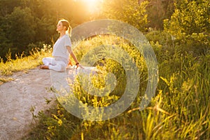 Happy woman sitting in lotus pose and doing bend in back outside in park on background of sunlight.