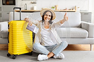 Happy Woman Sitting Near Suitcase, Holding Flight Tickets And Showing Thumb Up