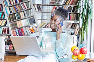 Happy woman sitting on sofa with laptop and talking on phone at home. Young successful businesswoman working from home while