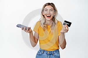 Happy woman with smartphone and credit card laughing, paying for something, winning money on phone, standing against