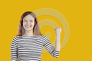 Happy woman smiles and points her thumb to copy space side on yellow studio background