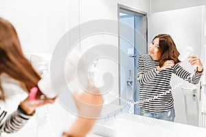 Happy woman smiling and drying her hair with hairdryer near the mirror in the bathroom