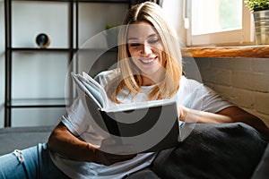 Happy woman smiling and reading book while sitting on sofa at home