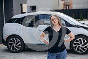 Happy woman standing near car, leaning elbow on windshield and putting her other hand on side door window. Girl