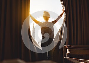 Happy woman stretches and opens curtains at window in morning