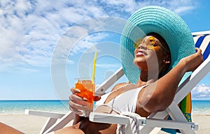 Happy woman is sunbathing on a beach deck chair, wearing sun hat and sunglasses, drinking a orange juice on a sunny day by the
