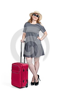 Happy woman in sunglasses, gray dress, hat and stiletto shoes stands with a red suitcase, isolated
