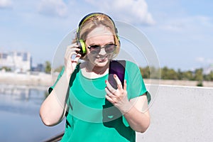 A happy woman in sunglasses and headphones taking a selfie photo on phone, chatting with friends on the street.