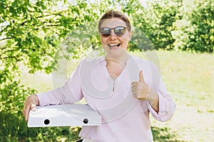 Happy Woman in sunglasses holding a box with pizza and showing thumbs up on nature background. Fast pizza delivery. Mock