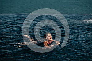 Happy woman in swimsuit swimming in infinity pool against seafront.