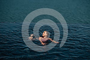 Happy woman in swimsuit swimming in infinity pool against seafront.