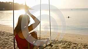 Happy woman on a swing by the ocean in the rays of the setting sun. Young woman is relaxing on the beach during vacation