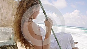 Happy woman swings near ocean, enjoys tropical vacation. Female in white dress leisurely swinging, breezy seaside