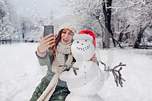 Happy woman takes selfie by snowman in Santa hat outdoors in snowy winter park. Christmas festive season
