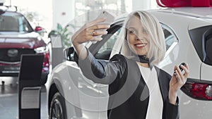 Happy woman taking selfies with keys to her new car at the dealership