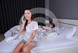 Happy woman talking on mobile phone in bed while her husband is