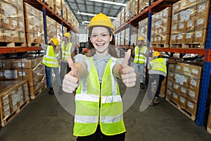 Happy woman thumbs up wearing safety uniform looking at camera standing in warehouse factory. Female professional worker stock