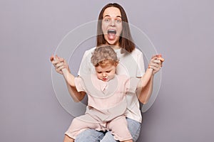 Happy woman with toddler child sitting sitting together isolated over gray background, mommy holding kid's hands
