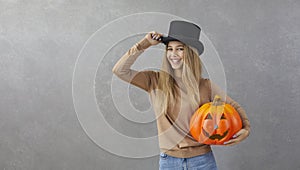 Happy woman in top hat holding Halloween pumpkin standing on grey copy space background