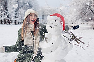 Happy woman toss snow up by snowman in Santa hat outdoors in snowy winter park. Christmas festive season