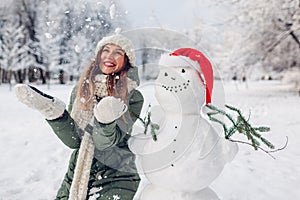 Happy woman tossing snow up by snowman in Santa hat outdoors in snowy winter park. Christmas festive season