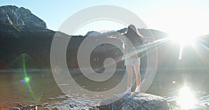 Happy woman tourist with a big backpack jumps on a large stone overlooking the lake and mountains, stands on the stone