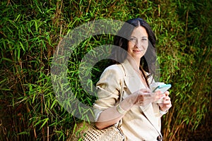 Happy woman traveller, texting on mobile phone and holding cofee cup, with green plant at background.