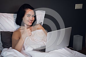 Happy woman typing on her laptop and showing thumbs up while lying in a bed at home