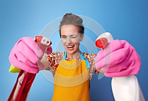 Happy woman using bottles of cleaning detergent as guns on blue