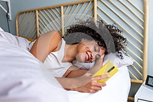 Happy woman using mobile phone in bed. Smiling black latina female looking mobile phone in the morning after waking up.