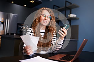 Happy woman using online banking app to pay bills, holding paychecks. Female book-keeping income, debts, loans, reciepts