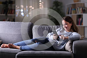 Happy woman using phone lying in the sofa at night at home