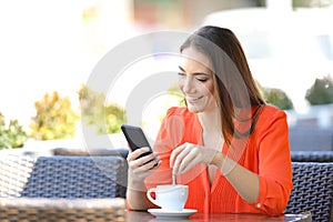 Happy woman using phone stirring coffee in a bar