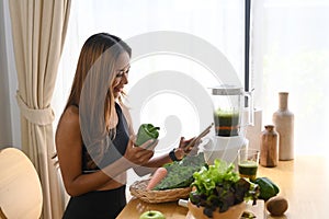 Happy woman using smart phone and making healthy green vegetable smoothie at home.