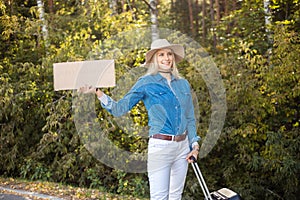Happy woman wait passing car in forest standing with suitcase and cardboard poster on roadside. Blonde lady escape from