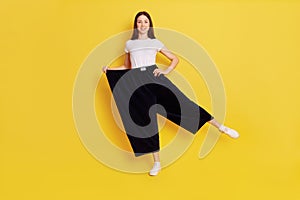 Happy woman after weight loss posing on one hand isolated over yellow background, wearing white casual t shirt and too big black