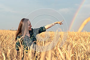Happy woman in wheat field with Golden spikelets pointing her finger at rainbow