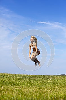 The happy woman in white bikini jumps in a summer green field against the blue sky