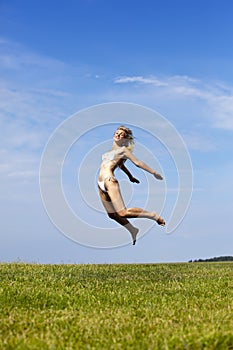 The happy woman in white bikini jumps in a summer green field against the blue sky