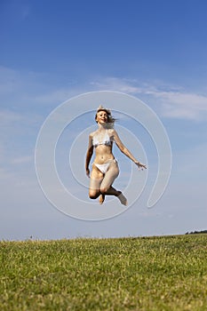 The happy woman in white bikini jumps in a summer green field against the blue sky