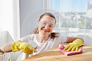 Happy woman in white shirt and yellow protective rubber gloves cleaning at home and wiping dust on wood table.