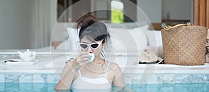 Happy woman in white swimsuit drinking hot tea in luxury swimming pool hotel, young female with hat enjoy in tropical resort.