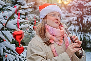 A happy woman in winter clothes plays the flute at the Christmas tree in a snow park on New Year Eve