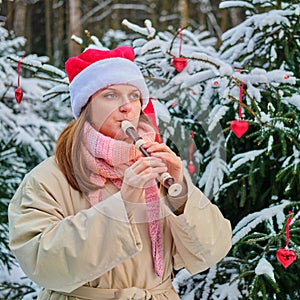 A happy woman in winter clothes plays the flute at the Christmas tree in a snow park on New Year Eve