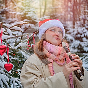 A happy woman in winter clothes plays the flute at the Christmas tree in a snow park on New Year Eve