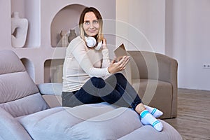 Happy woman in wireless headphones with phone sits on sofa indoors.