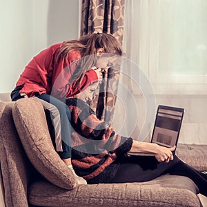 Happy woman working at home on computer and cute kid play with mother. Sitting on sofa, daughter covering mother the eyes with