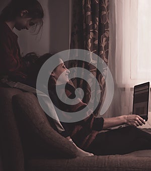 Happy woman working at home on computer and fun kid playing with mother. Sitting together on the sofa with smiling playful faces.