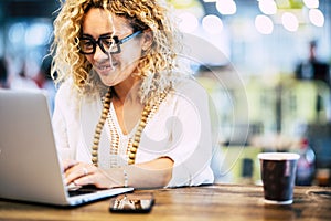 Happy woman working on laptop at coffee shop. Woman in eyeglasses and beads necklace using laptop on desk with mobile phone and