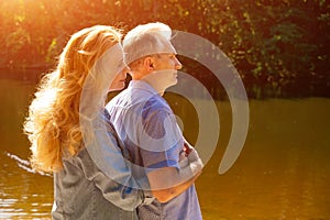 A happy woman embraces a man behind her back, standing on the shore of the lake, in the rays of the sun at sunset